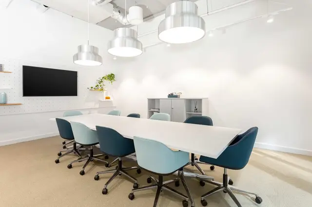 Meeting room with a long white table, blue chairs, pendant lights and a wall-mounted screen.